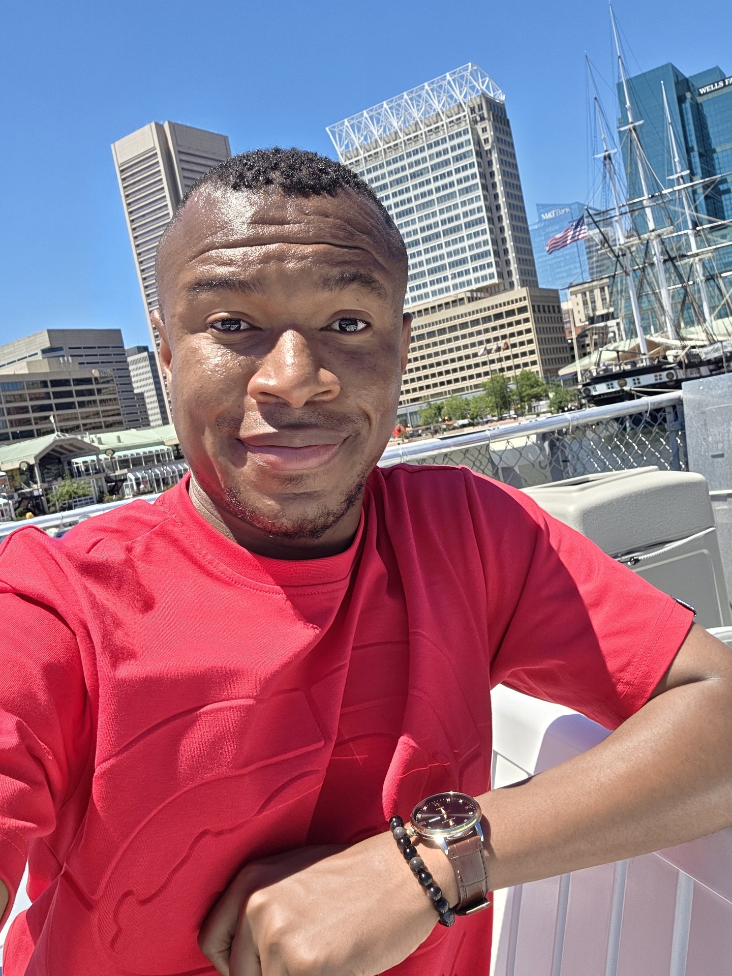 Kingsley seated beside the waterfront in a red shirt with city buildings in the distance